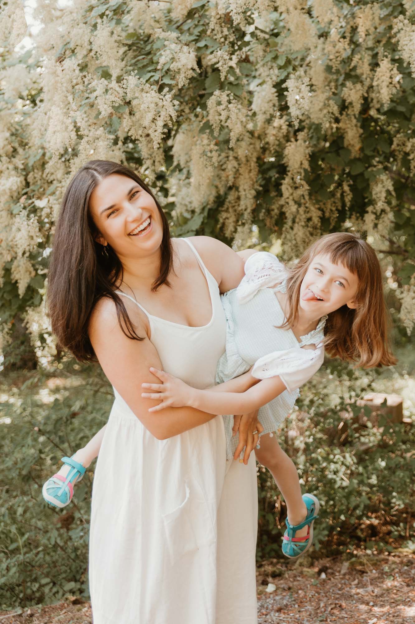 A little girl with a big smile is being swung by her mom. Both are looking at the camera, with the mom holding her daughter close. They are outdoors, with greenery in the background, and both are wearing casual, comfortable clothes.