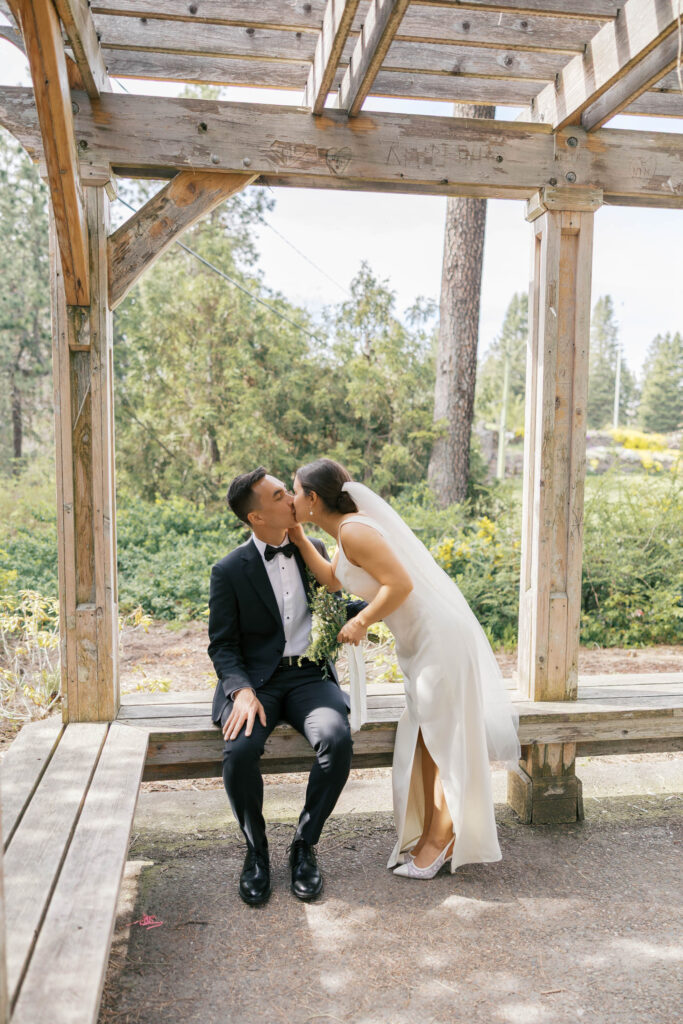 A bride is standing and leaning over to kiss the sitting groom in a gazebo at Manito Park in Spokane. 