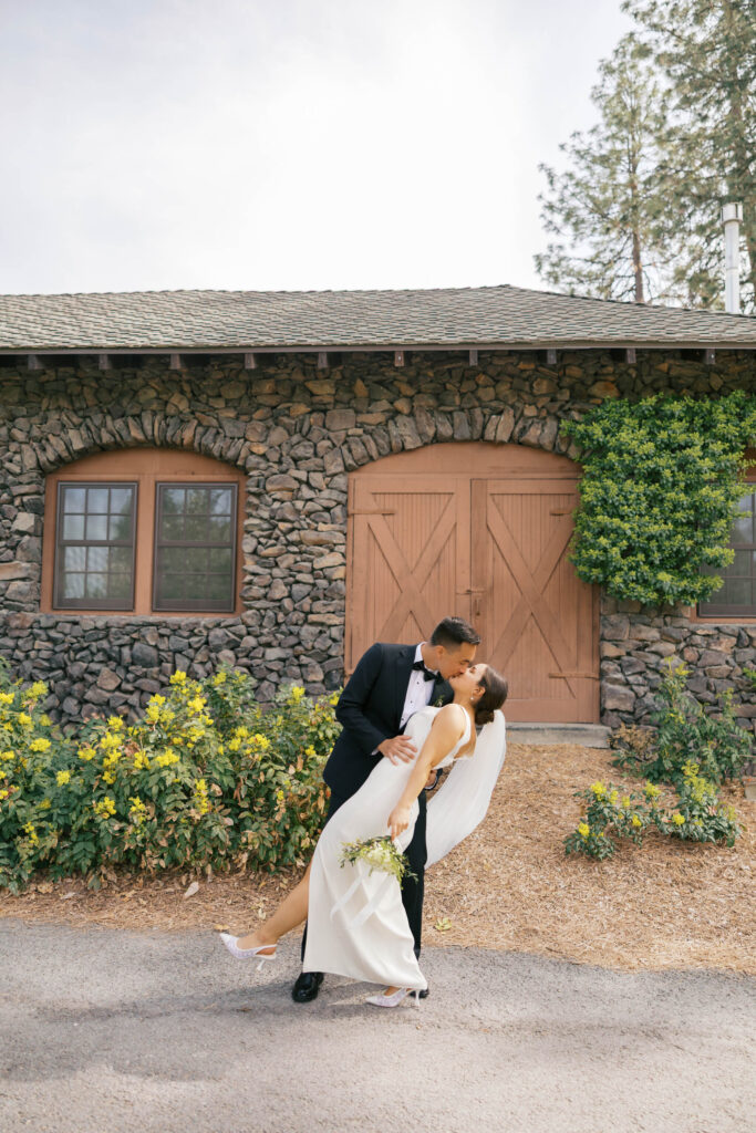 A groom is dipping the bride in front of a cobblestone building in Manito Park. 