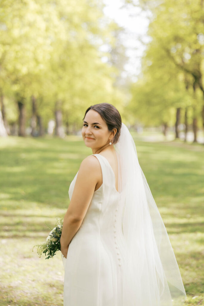 A bride looking over her shoulder smiling, she is holding flowers and wearing her veil. 