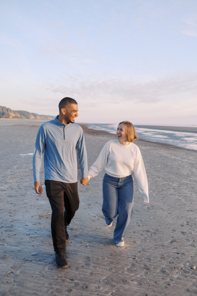Julia and A'lon walking hand-in-hand and smiling at each other on the sand