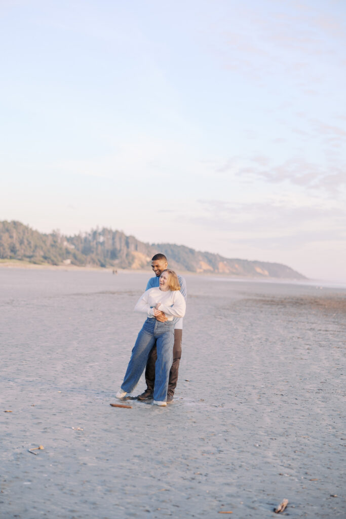 A'lon hugging Julia from behind, both laughing on a wide-open beach