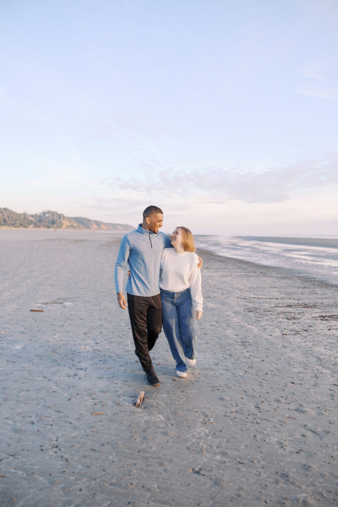 Engaged couple walking arm-in-arm along the shoreline as the tide rolls out