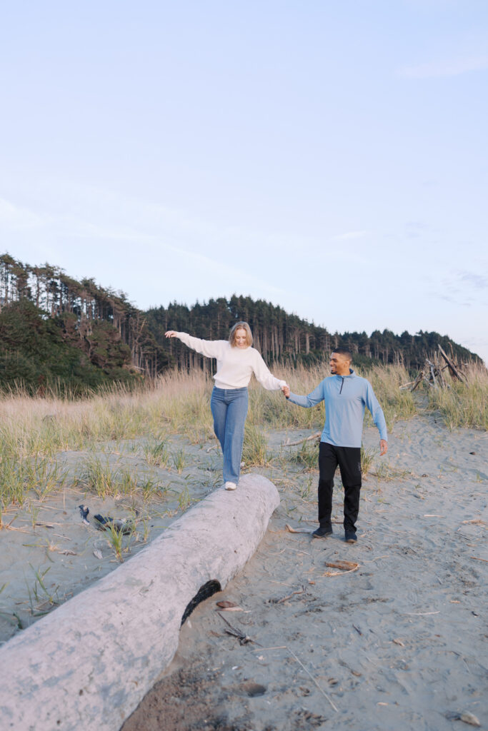 Julia balancing on driftwood while A'lon holds her hand during a Seabrook beach engagement session
