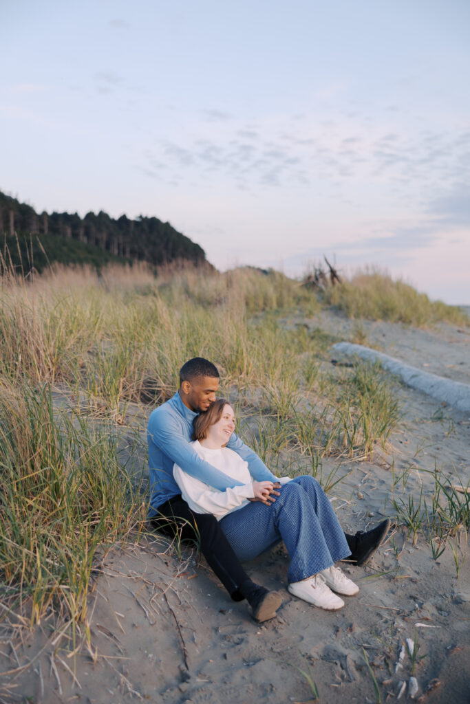 Engaged couple sitting close in the dune grass, smiling out toward the water